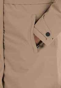 Lichtbeige jas met een slank ontwerp, zachte textuur, zijzak met rits en een knoopdetail op de manchetten.