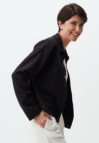 Zwarte button-up jas met een grote borstzak, gemaakt van zachte stof, met lange mouwen en een ontspannen pasvorm. Draagt over een wit kledingstuk.