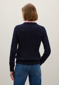 Marineblauer Kabelstrickpullover mit geripptem Saum und Bündchen, ausgestattet mit einem klassischen Rundhalsausschnitt. Kombiniert mit blauen Jeans, die detaillierte Nähte zeigen.