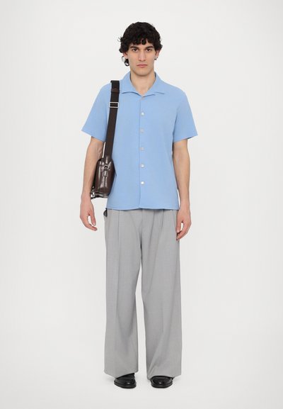 Camicia azzurra a maniche corte con bottoni, pantaloni grigi a gamba larga e scarpe nere. Borsa marrone appesa alla spalla.