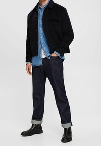 Černá vlněná bunda přes světle modrou džínovou košili, v kombinaci s tmavě modrými džínami s vyhrnutými lemy a černými koženými kotníkovými botami.