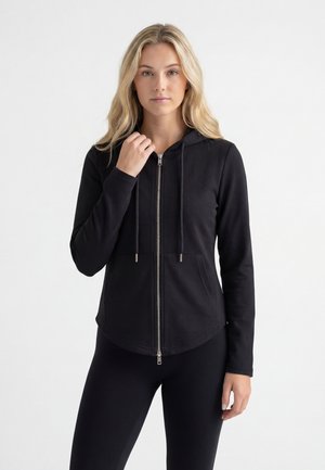 Jeune femme aux longs cheveux blonds portant un sweat à capuche noir zippé avec poches avant et un legging noir sur un fond blanc uni.