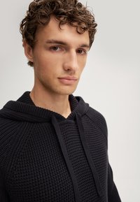 Czarna dzianinowa bluza z kapturem z teksturowanym wzorem, z regulowanym kapturem i rękawami raglanowymi. Zbliżenie na górną przednią część odzieży.