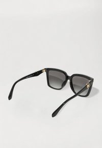 Ochelari de soare din plastic negru cu rame largi, lentile gri cu gradient și accente aurii pe tâmple, având un design geometric.