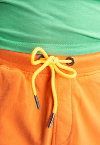 Oranje sweatpants met een geribbeld tailleband, voorzien van een gele trekkoord met zilveren metalen uiteinden, gedragen over een groene t-shirt.
