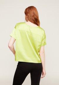 Limetkově zelený saténový top s krátkým rukávem, volným střihem, kulatým výstřihem a knoflíkovým detailem na zádech. Spárováno s černými kalhotami.
