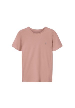Joven con el cabello hasta los hombros que lleva una camiseta malva, pantalones cortos beige texturizados y sandalias marrones sin cierre, de pie frente a un fondo liso.