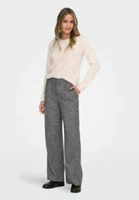 Moteris stovi su ranka kišenėje, vilkėdama baltą ilgasvarkę, pilkas plačias kelnes ir tamsiai rudus loafers batus ant baltos fono.