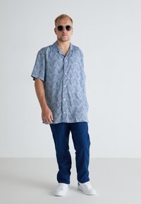 Chemise à manches courtes, à motif bleu, en tissu léger, associée à un pantalon bleu foncé et des baskets blanches. Accessoirisée avec des lunettes de soleil.