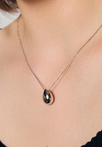Błyszczący srebrny wisior w kształcie kropli wiszący na delikatnym łańcuszku, spoczywający na gładkiej, lekko piegowatej skórze. Częściowo widoczne czarne ubranie.