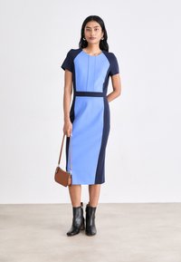 Μπλε φόρεμα με χρωματικές μπλοκ, κοντά μανίκια, με ναυτικές λεπτομέρειες και εφαρμοστό περίγραμμα. Συνδυασμένο με μαύρες μπότες αστράγαλου και καφέ τσάντα.