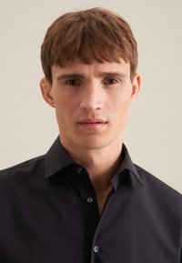 Zwart overhemds met een spitse kraag, knoopsluiting en gladde stof. Subtiele glans en gestructureerde pasvorm versterken de formele uitstraling.