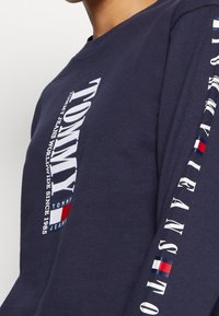 Námořnická košile s dlouhým rukávem vyrobená z bavlny, na přední straně a rukávu s bílým nápisem "TOMMY". Obsahuje červené, bílé a modré logo akcenty.