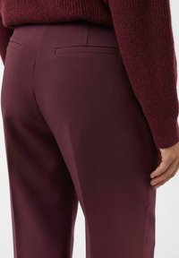 Person wearing dark burgundy tailored trousers and a matching knitted sweater, shown from the waist down with one hand visible.