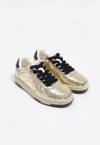 Zapatillas doradas con un acabado texturizado, cordones negros y un interior crema. Presentan una punta redonda y una suela negra.