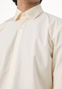 Gros plan d'une personne portant une chemise crème boutonnée avec col, montrant le col et le haut du torse.