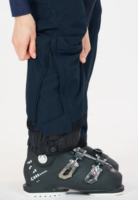 Botas de esquí negras combinadas con pantalones azul marino que tienen un bolsillo con cremallera, puños elásticos y un acabado texturizado para mayor durabilidad.