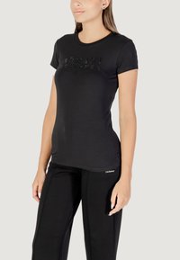 Camiseta de algodón negra con cuello redondo, mangas cortas y detalle de logo en relieve en la parte frontal. Combinada con pantalones negros.