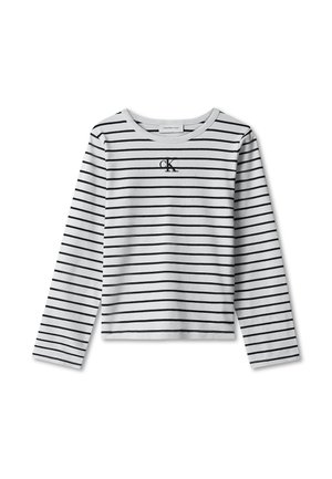 Kind met krullend haar dat een zwart-wit gestreept shirt met lange mouwen draagt, lichtblauwe wijde jeans, zwarte riem en zwarte schoenen, staand tegen een grijze achtergrond.