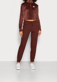 Kvinna klädd i en brun Nike kortärmad zip-up sweatshirt och matchande joggers med vita sneakers, stående mot en enkel ljus bakgrund.