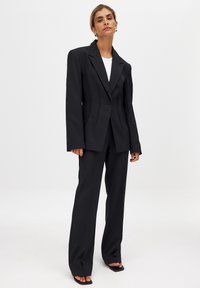 Black tailored suit featuring a structured blazer with a single button, combined with straight-leg trousers, offering a matte texture and sharp lines.