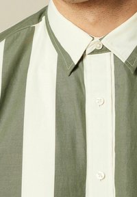Hedvábně zeleno-krémová pruhovaná košile s límečkem, která má jemnou texturu a viditelné švy podél švů.