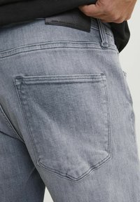 Pilkos denimo džinsai su lygiu paviršiumi, turi galinį kišenę ir juostos dalyje užrašytą juodą odinį etiketę su įspaustu "JACK & JONES".