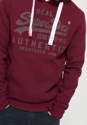 Bordeauxroter Hoodie mit weißen Kordeln, vorderer Kängurutasche und dem Schriftzug "Real Superdry Authentic Registered JPN" in dunklem Druck.