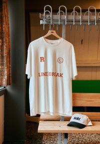 T-shirt bianca in cotone con grafiche rosse: "R", uno stemma e "LINE|BREAK." Appesa a una panchina di legno con un cappellino grigio e nero accanto.
