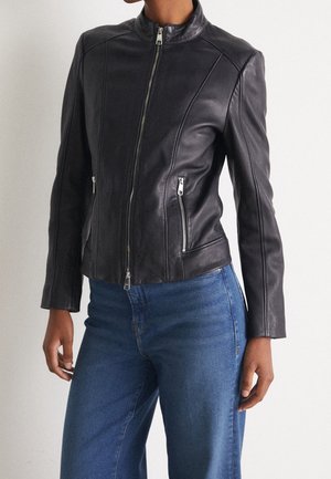 Vrouw die een nauwsluitend zwart leren jack met zilveren ritsen draagt, gecombineerd met een hoog Taille blauwe jeans tegen een effen achtergrond.