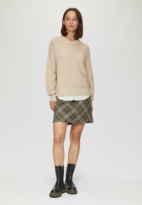 Helle beigefarbene Strickjacke mit horizontalen Streifen, über einem weißen Hemd getragen. Olivgrüner kariert Rock und schwarze klobige Knöchelstiefel.