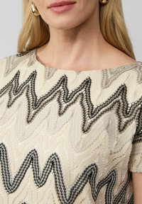Beige top met zigzagpatroon in zwart en taupe, met een brede halslijn en korte mouwen. De textuur is geweven met borduurdetails.