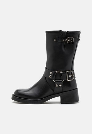 Botte noire en cuir mi-mollet avec un petit talon, deux boucles argentées réglables et un détail décoratif en forme d'anneau sur le côté.
