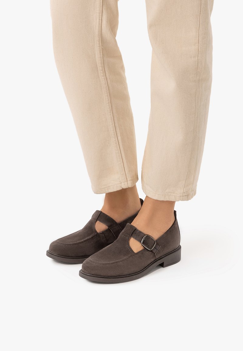 Personne portant un pantalon beige et des chaussures style Mary Jane en suède marron foncé avec une boucle latérale, debout sur un fond blanc uni.