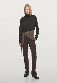 Dunkelgrüner Rollkragenpullover, mit einem braunen Wickelakzent an der Taille, kombiniert mit dunkelbraunen Hosen und schwarzen Schuhen. Einfaches, strukturiertes Design.