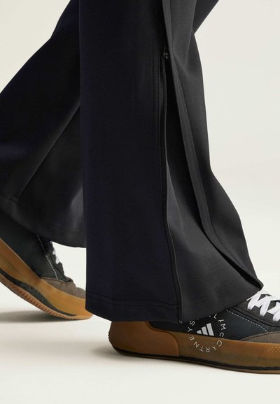 Schwarze Sporthose mit seitlichen Reißverschlüssen, kombiniert mit tief geschnittenen Turnschuhen in Schwarz und Beige mit strukturierten Gummisohlen.