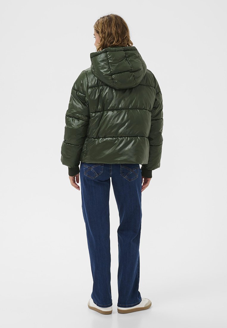 Puffer-Jacke in glänzendem Dunkelgrün mit horizontalen Nähten und Kapuze, kombiniert mit blauen Jeans und weißen Turnschuhen, von hinten gesehen.