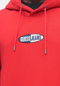 Κόκκινο βαμβακερό hoodie με μπροστινή τσέπη καγκουρό, κουκούλα με κορδόνια και κεντημένο λογότυπο "GUESS JEANS" στο στήθος· ομαλή υφή και casual σχεδιασμός.