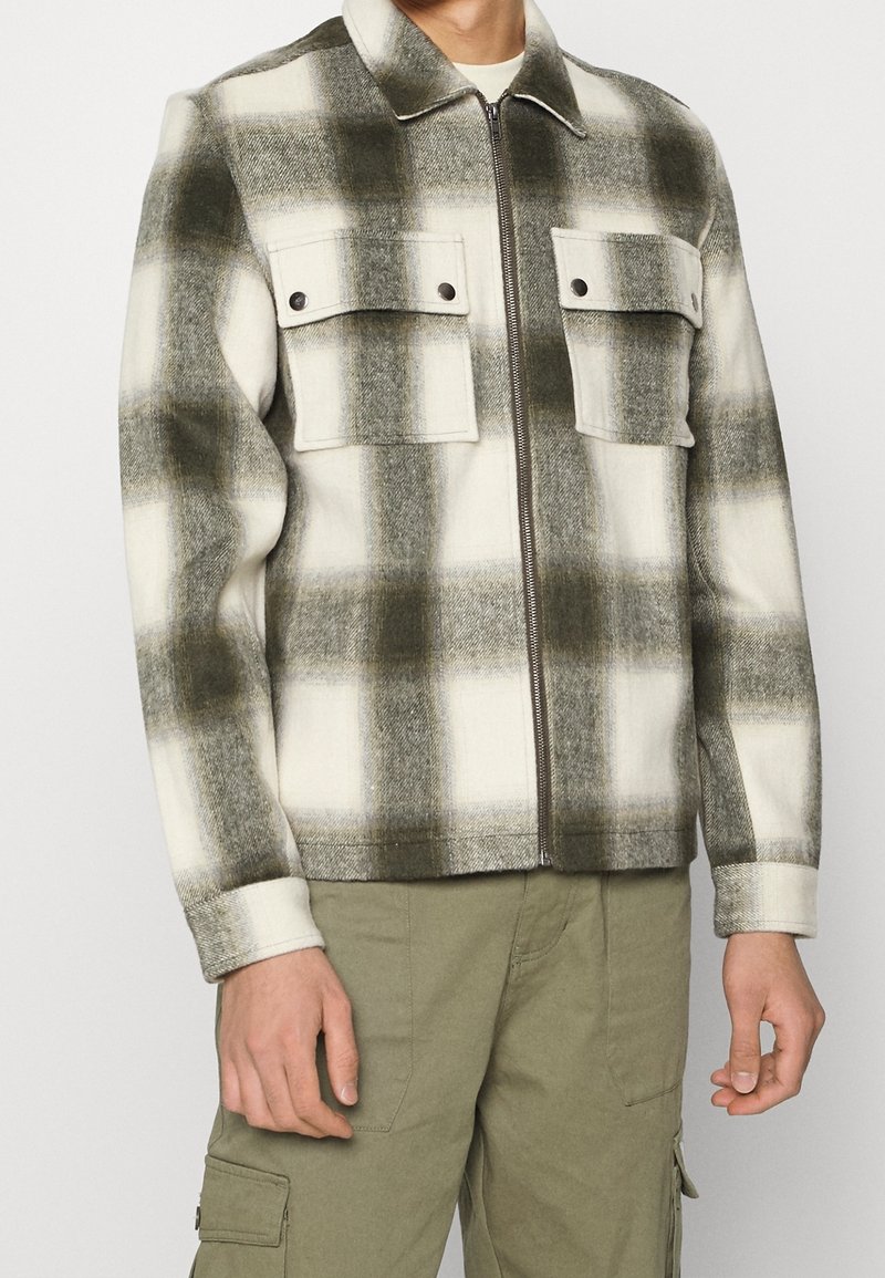 Chaqueta de cuadros en tonos crema y verde, con dos bolsillos en el pecho, cierre de cremallera y un diseño con cuello de tejido texturizado.