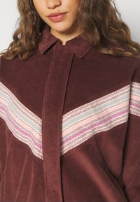 Vrouw in een bordeauxrode corduroy jas met een chevronpatroon van golvende strepen in roze, wit, oranje en blauw over de borst.