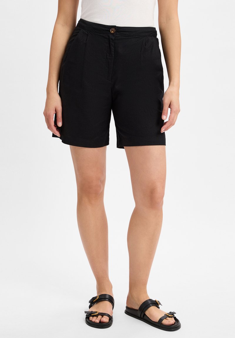 Pantaloni neri con chiusura a bottone, dotati di tasche laterali e un tessuto leggermente testurizzato. Abbinati a sandali neri piatti.