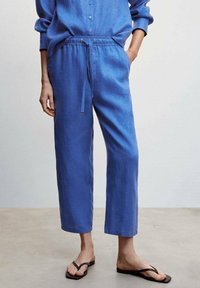 Blauwe linnen broek met een tailleband met trekkoord, wijde cropped pijpen en een losse pasvorm. Draagt zwarte teenslippers waarmee blote voeten zichtbaar zijn.