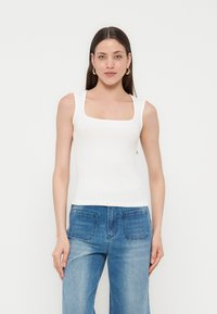 Γυναίκα που φοράει λευκό αμάνικο τοπ με τετράγωνη λαιμόκοψη και μπλε τζιν, στέκεται μπροστά σε ένα απλό φωτεινό φόντο.