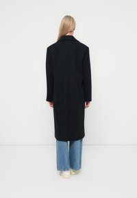 Abrigo largo negro de lana, con una silueta estructurada, solapa muesca y mangas largas, combinado con pantalones vaqueros holgados y zapatillas.