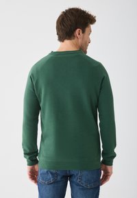 Sweat-shirt vert foncé à manches longues et col rond. Présente une texture lisse et des poignets côtelés, associé à un jean en denim bleu.