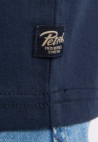Tissu en coton bleu marine avec une texture à côtes, comportant une étiquette tissée rectangulaire avec "Petrol Industries" en relief dans une couleur crème.