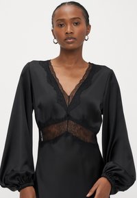 Melna kleita ar garām balona piedurknēm, krūšu daļā caurspīdīgs mežģīņu ielikums un sarežģīta mežģīņu apdare gar kakla izgriezumu. Gluda auduma tekstūra.