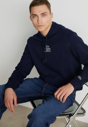 Sudadera azul marino de mezcla de algodón, con bolsillo tipo canguro, capucha con cordón y logo bordado. Combinada con jeans de mezclilla.