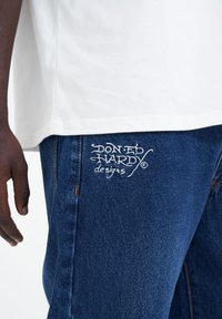 Jeans de denim en azul oscuro con costuras doradas, que presentan el logotipo bordado en blanco "DON ED HARDY designs" en el muslo izquierdo. Mezcla de algodón suave.