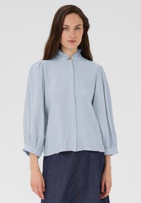 Blouse bleu clair avec un col à volants haut, des manches structurées et une fermeture à boutons. Tissu doux avec un léger éclat, associée à une jupe en denim foncé.